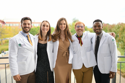 Five people standing outdoors, arms around each other, wearing the white coats symbolic of the medical profession