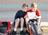 Two boys with backpacks sit on sidewalk and go over homework