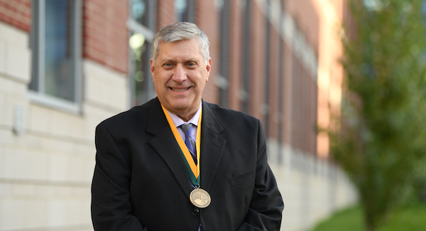 Randall Holcombe in a black suit, grey tie, white shirt and wearing a gold medallion with gold and green ribbon