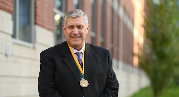 Holcombe_Invest_600x325 Randall Holcombe in a black suit, grey tie, white shirt and wearing a gold medallion with gold and green ribbon