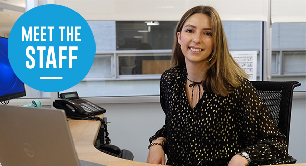 Photo of woman smiling wearing a black dress, sitting at her desk with a blue sticker that says 'meet the staff'