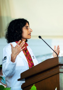 Devika Singh standing at a podium in a white coat speaking