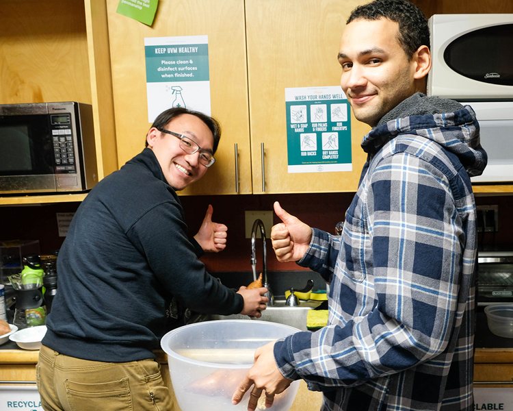 Two people standing next to a sink, one washing a sweet potato, the other holding a bowl of washed sweet potatoes, both smiling at the camera and giving a 