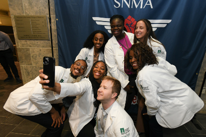 Seven people posing for a selfie photo in front of a blue, white, and red banner with the logo of the Student National Medical Association